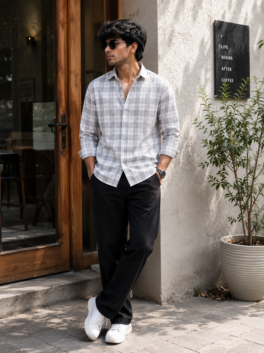 Man wearing a checkered shirt and black pants standing outside a building with a plant in the background.
