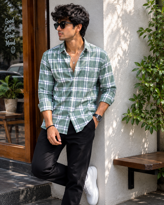 Man wearing a green plaid shirt and sunglasses leaning against a wall with plants around.