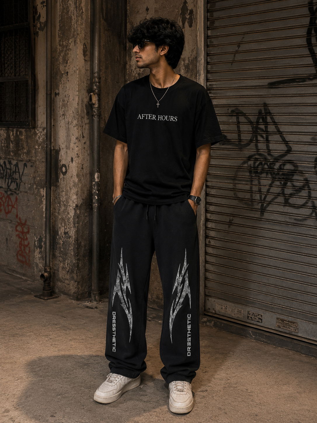 Person wearing a black 'AFTER HOURS' t-shirt and black pants with white designs, standing in an urban setting.
