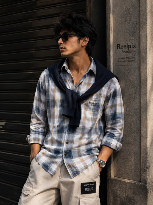Man wearing a plaid shirt and sunglasses leaning against a wall.
