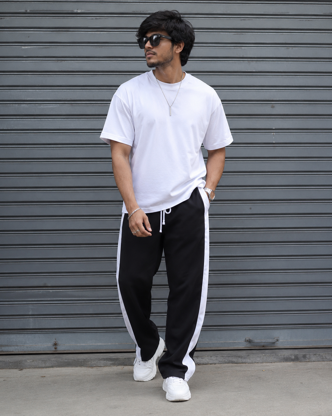 Man wearing a white t-shirt and black pants with white stripes standing in front of a gray metal shutter.