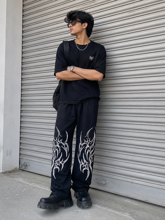 Person wearing black clothing with tribal designs, standing against a metallic shutter.